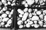 A collection of potatoes with dirt on their surfaces is arranged in crates, displaying a variety of sizes and round to oval shapes. The image is in black and white, emphasizing the textures and contrasts of the potatoes and the crates.