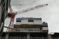 A large construction site with a tall red crane towering over a partially constructed building. The building features multiple levels with metal and wooden frameworks. The sky is overcast, and there are wires and protective barriers evident on the structure. Workers can also be seen on the structure.