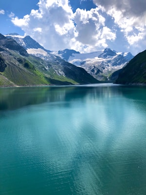 A serene mountain landscape with a clear turquoise lake reflecting the surrounding snow-capped peaks and lush green hillsides under a partly cloudy blue sky.