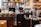 A cozy café setting features multiple coffee grinders filled with coffee beans, surrounded by stacked coffee cups and various equipment. Shelves in the background display an assortment of jars and bottles.
