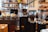 A cozy café setting features multiple coffee grinders filled with coffee beans, surrounded by stacked coffee cups and various equipment. Shelves in the background display an assortment of jars and bottles.