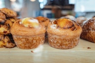 Two pastries with golden-brown, flaky layers and a caramelized top, set against a backdrop of additional baked goods on a wooden surface.