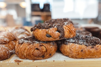 A pile of freshly baked pastries topped with powdered sugar and chocolate, placed on a wooden surface. The pastries have a golden-brown crust and are layered with visible swirls, indicating a flaky texture.