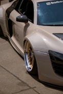 A luxury sports car with a sleek design is parked on a concrete surface. The car features distinctive gold alloy wheels and a light beige exterior. The side mirror and part of the windshield are visible, adorned with branding and a small decal. Shadows cast by the sun are evident on the smooth surface of the vehicle.