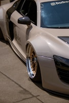 A luxury sports car with a sleek design is parked on a concrete surface. The car features distinctive gold alloy wheels and a light beige exterior. The side mirror and part of the windshield are visible, adorned with branding and a small decal. Shadows cast by the sun are evident on the smooth surface of the vehicle.