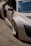 A luxury sports car with a sleek design is parked on a concrete surface. The car features distinctive gold alloy wheels and a light beige exterior. The side mirror and part of the windshield are visible, adorned with branding and a small decal. Shadows cast by the sun are evident on the smooth surface of the vehicle.