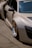 A luxury sports car with a sleek design is parked on a concrete surface. The car features distinctive gold alloy wheels and a light beige exterior. The side mirror and part of the windshield are visible, adorned with branding and a small decal. Shadows cast by the sun are evident on the smooth surface of the vehicle.