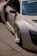 A luxury sports car with a sleek design is parked on a concrete surface. The car features distinctive gold alloy wheels and a light beige exterior. The side mirror and part of the windshield are visible, adorned with branding and a small decal. Shadows cast by the sun are evident on the smooth surface of the vehicle.