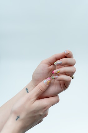 Two hands are shown with the focus on colorful, artistic nail designs. Each nail has a unique pattern with abstract shapes and multiple colors. The background is a soft, light color, adding emphasis to the vibrant nail art. Tattoos are visible on the wrists and fingers of the hands.