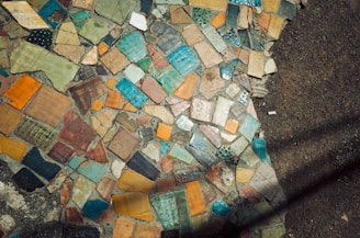 A mosaic of multicolored tiles in various shapes and sizes is embedded in a gritty, dark concrete surface. The tiles exhibit a vibrant mix of hues including blue, orange, green, and brown, arranged in an abstract, patchwork pattern.