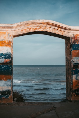 An old, weathered archway frames a view of the ocean, with waves gently rolling towards the shore. The archway is painted in faded hues of blue, orange, and beige, suggesting a connection with the sea and sky beyond.