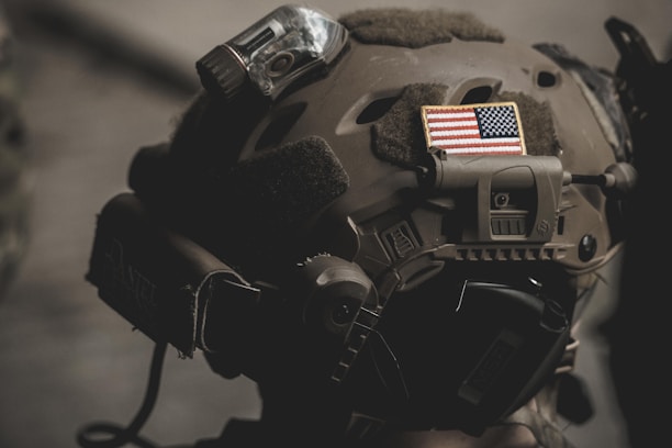 A close-up of a tactical helmet adorned with an American flag patch, featuring attachments like a camera or light. The helmet appears military-grade, with muted earth tones and practical design elements.