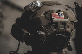 A close-up of a tactical helmet adorned with an American flag patch, featuring attachments like a camera or light. The helmet appears military-grade, with muted earth tones and practical design elements.