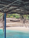 A scenic beach view is partially framed by a rustic, thatched roof. The crystal-clear turquoise water meets a sandy shore, and people can be seen relaxing on the beach. In the background, a cliff displays the word 'ARRAFAL', and there is some greenery atop the cliff.