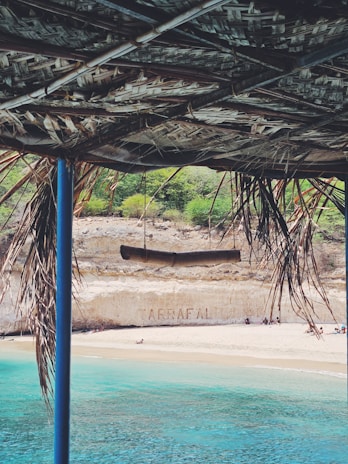 A scenic beach view is partially framed by a rustic, thatched roof. The crystal-clear turquoise water meets a sandy shore, and people can be seen relaxing on the beach. In the background, a cliff displays the word 'ARRAFAL', and there is some greenery atop the cliff.