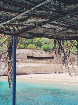 A scenic beach view is partially framed by a rustic, thatched roof. The crystal-clear turquoise water meets a sandy shore, and people can be seen relaxing on the beach. In the background, a cliff displays the word 'ARRAFAL', and there is some greenery atop the cliff.