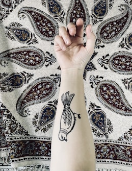 A person&rsquo;s forearm is extended in front of an intricately patterned textile. The arm features a tattoo of an upside-down bird, drawn in a minimalistic black ink style. The background consists of a paisley design with rich colors such as maroon, blue, and beige.
