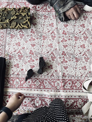 A decorative textile with intricate red and white patterns fills the background. On the top left, a detailed wooden block with carved floral designs is placed. Two small black bird figurines rest near the center. Hands and part of a garment can be seen on the right side, suggesting interaction with the objects.