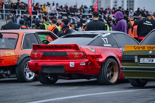 Numerous people stand in a crowd near a racetrack, observing a lineup of tuning automobiles. There are several modified cars, including a red one with the label 'Rocket Bunny' and a number '17'. Bright orange safety vests are visible on some individuals, suggesting event staff or security personnel.