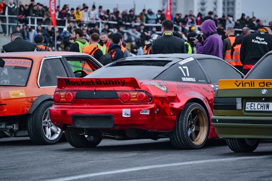 Numerous people stand in a crowd near a racetrack, observing a lineup of tuning automobiles. There are several modified cars, including a red one with the label 'Rocket Bunny' and a number '17'. Bright orange safety vests are visible on some individuals, suggesting event staff or security personnel.