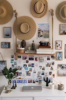 A decorative wall features an arrangement of straw hats, framed photographs, and postcards. A desk below holds a laptop, a potted plant, and various decorative items, including a small Eiffel Tower and a California license plate. Books and a vintage camera are placed on a wooden shelf above the desk.