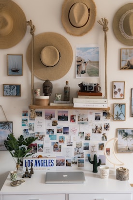 A decorative wall features an arrangement of straw hats, framed photographs, and postcards. A desk below holds a laptop, a potted plant, and various decorative items, including a small Eiffel Tower and a California license plate. Books and a vintage camera are placed on a wooden shelf above the desk.