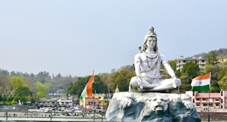 A large statue of a meditating deity, adorned with traditional ornaments, is set against a scenic background with lush trees and distant hills. Two Indian flags flank the statue, emphasizing a cultural and national theme. Buildings are visible in the background, suggesting a populated area nearby.
