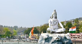 A large statue of a meditating deity, adorned with traditional ornaments, is set against a scenic background with lush trees and distant hills. Two Indian flags flank the statue, emphasizing a cultural and national theme. Buildings are visible in the background, suggesting a populated area nearby.