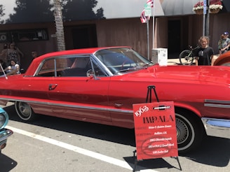 A bright red 1963 Chevrolet Impala is parked on a street with a signboard in front, providing details about the car. Several people are around the car, some sitting on a bench under the shade of a tree. There's a bicycle in the background near another person standing. The setting appears to be a car show or exhibition.