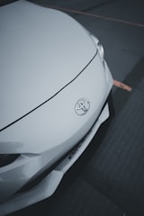 A close-up of the front of a white car featuring a Toyota emblem. The smooth, sleek design of the car's body is highlighted, with a visible headlight and part of the bumper.