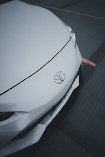 A close-up of the front of a white car featuring a Toyota emblem. The smooth, sleek design of the car's body is highlighted, with a visible headlight and part of the bumper.
