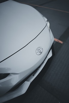 A close-up of the front of a white car featuring a Toyota emblem. The smooth, sleek design of the car's body is highlighted, with a visible headlight and part of the bumper.