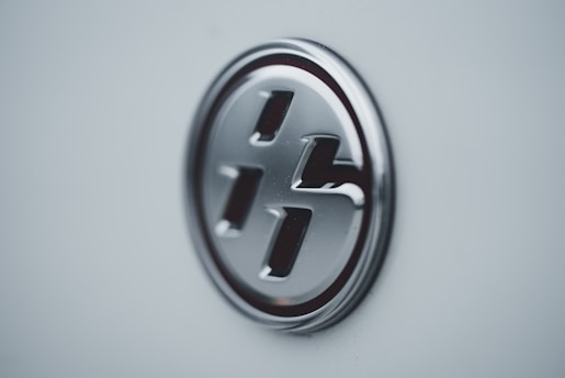 A close-up of a metallic, circular emblem on a light-colored surface, featuring a stylized letter design.