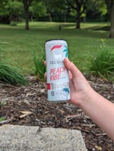 A hand holds a can of Celsius Peach Vibe energy drink in an outdoor setting with a grassy area and trees in the background.