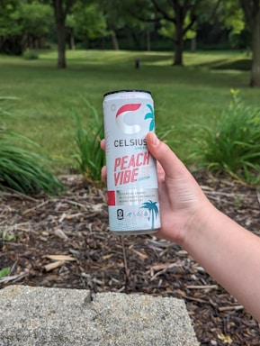 A hand holds a can of Celsius Peach Vibe energy drink in an outdoor setting with a grassy area and trees in the background.
