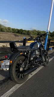 A motorcycle is parked on the side of a road, with lush green trees and a clear blue sky in the background. The road is bordered by a railing, and the area appears open and serene.