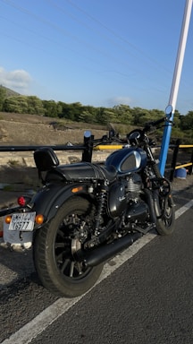 A motorcycle is parked on the side of a road, with lush green trees and a clear blue sky in the background. The road is bordered by a railing, and the area appears open and serene.