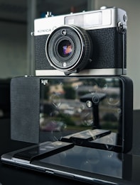 A vintage Konica camera is perched on top of a modern multi-lens digital camera. The reflection of the vintage camera is visible on the digital camera's surface. The setup rests on a dark, glossy surface with an indistinct background.
