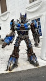 A large robot with intricate mechanical details stands in front of a white building. The robot features blue and metallic colors with components resembling gears and bolts. It has a humanoid shape and appears to be in a static position.