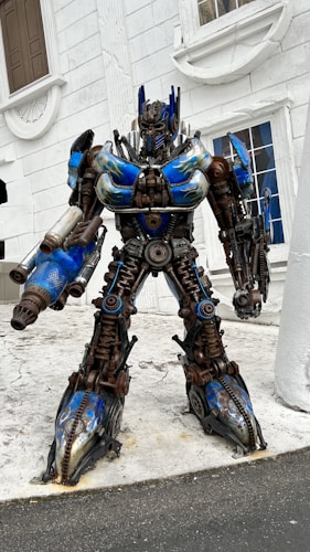 A large robot with intricate mechanical details stands in front of a white building. The robot features blue and metallic colors with components resembling gears and bolts. It has a humanoid shape and appears to be in a static position.