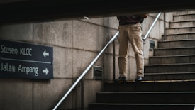A person stands on a stairway next to a wall with directional signs pointing towards Stesen KLCC and Jalan Ampang. The person is dressed in light-colored pants and dark shoes, partially obscured by the structural frame.