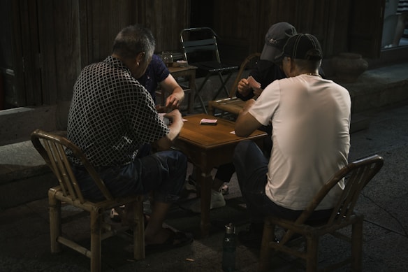 Three men are seated around a small wooden table playing cards. They are outdoors, and the setting is dimly lit, suggesting a night-time environment. Two men are sitting on wooden chairs, and another is seated on a metal folding chair. One of the men is wearing a patterned shirt and another a plain white t-shirt.