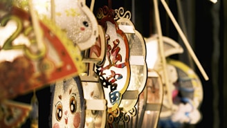 A collection of decorative paper lanterns with intricate patterns and cartoon faces, featuring vibrant colors and traditional Chinese characters. The lanterns hang from thin poles and appear illuminated from within, casting a warm glow.