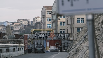 Several multi-story buildings with a gated entrance labeled 'Seaside Langtai' are visible. A starfish decorates the gate, and a black car is parked in front. The scene includes rocky terrain and a blurred sign in the foreground.