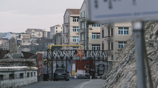 Several multi-story buildings with a gated entrance labeled 'Seaside Langtai' are visible. A starfish decorates the gate, and a black car is parked in front. The scene includes rocky terrain and a blurred sign in the foreground.
