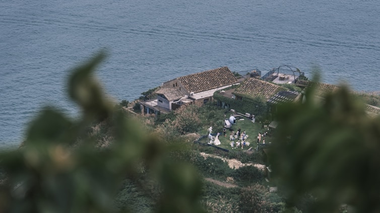 A scenic view features a small rustic building with a tiled roof, surrounded by greenery near a large body of water. People are gathered outside, possibly at a small event or gathering. The scene is partially framed by blurred foliage in the foreground.
