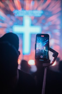 A large illuminated cross stands prominently in a vibrant background with colorful light streaks radiating outward. In the foreground, a person is holding a smartphone with a stabilizer, capturing a video of the scene.