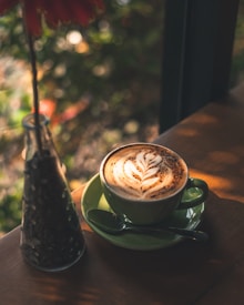 A cup of coffee with a beautifully crafted latte art design sits on a green saucer with a spoon placed beside it. The cup is on a wooden surface and next to a bottle containing coffee beans. The background is softly blurred, with hints of greenery and soft lighting creating a warm and cozy ambiance.