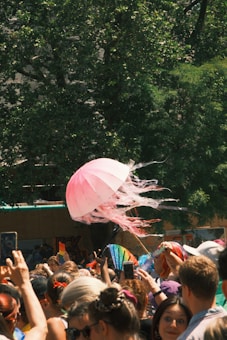 A vibrant crowd gathers outdoors, with many people holding colorful objects such as a pink jellyfish-shaped umbrella, rainbow flags, and fans. The scene is lively, with sunlight streaming through the trees in the background.
