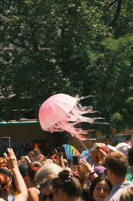 A vibrant crowd gathers outdoors, with many people holding colorful objects such as a pink jellyfish-shaped umbrella, rainbow flags, and fans. The scene is lively, with sunlight streaming through the trees in the background.